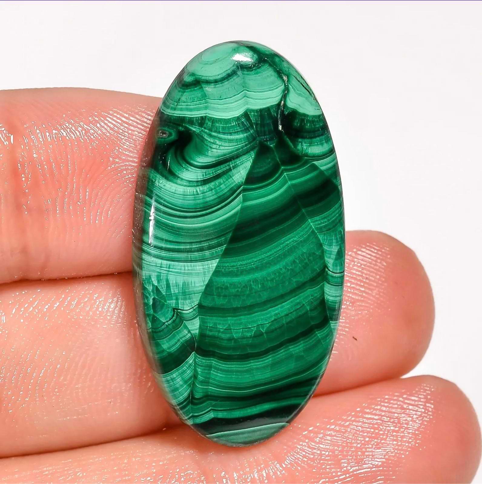 Polished oval Malachite stone with rich green banding held between fingers, used for Taurus zodiac energy, against a white background.